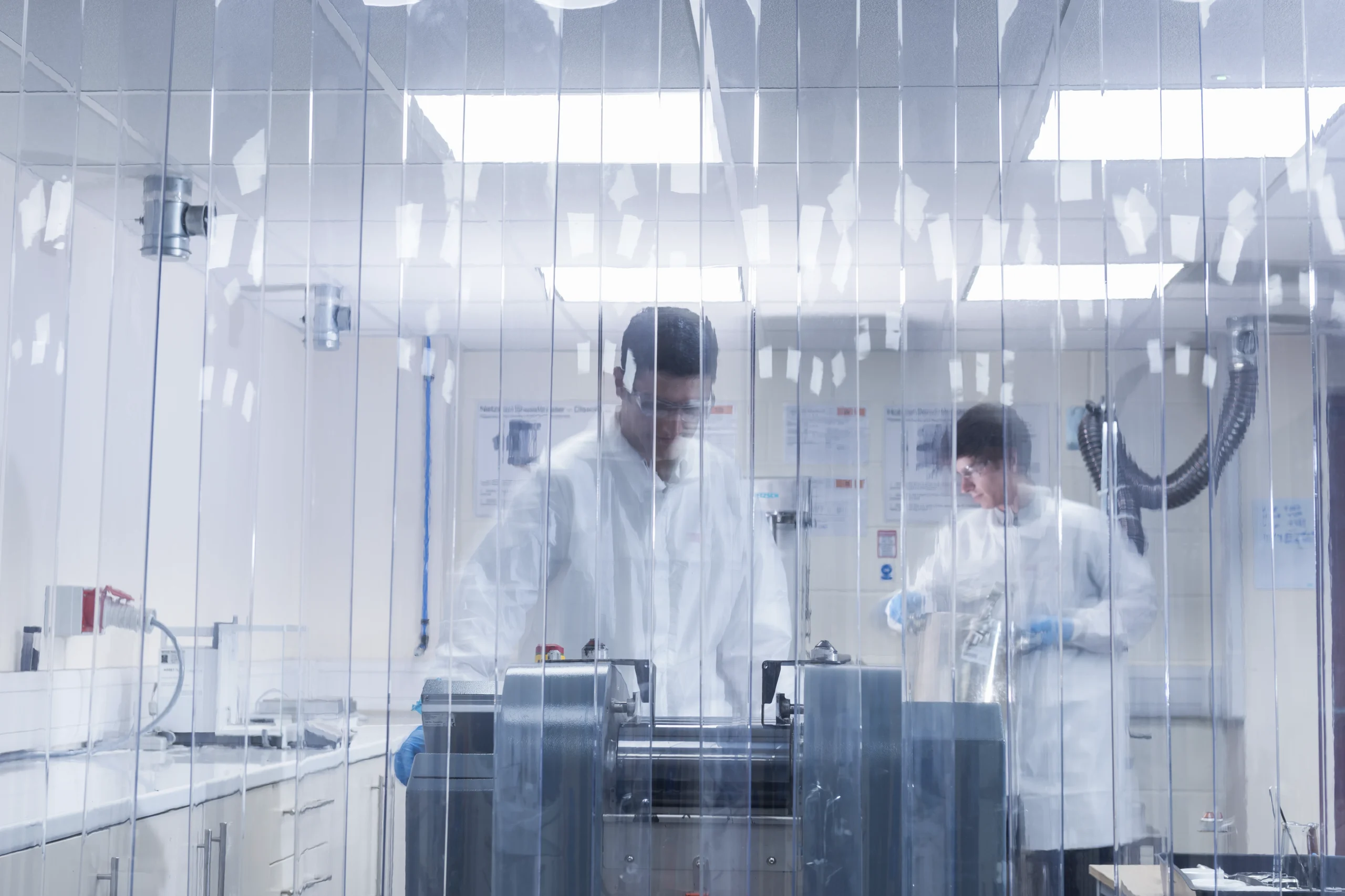 Technicians in lab coats working with machinery behind protective strip curtains in a controlled laboratory environment